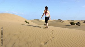 Young woman walks barefoot on sand dunes with sea views on the background in Maspalomas, Gran Canaria. Lonely lady exploring desert in Canary Islands, Spain. Travel adventure, summer holidays concept