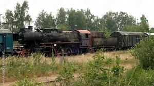 An old rusty black steam locomotive with a steam propulsion system and cars on abandoned tracks.