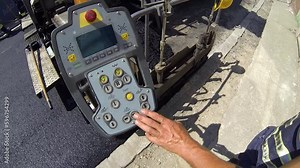 Road Construction Worker Operates the Asphalt Paver Machine. Road Construction Industry. Close-up of the Asphalt Paver's Control Panel. Worker's Hand at the Asphalt Paver's Console. Asphalt Paving.