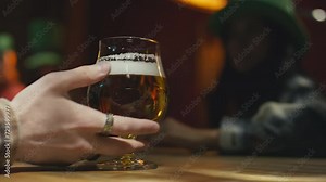 Closeup of unrecognizable bartenders hand serving glass of filtered beer, ale or lager to unidentified female customer sitting at bar in green hat celebrating St Patricks Day