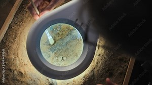 Close up of paleoanthropologist or archaeologist cleaning bone of ancient prehistoric human with brush. Scientist works with fossil remains in archaeological laboratory. View through magnifying lamp.