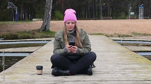 Woman in hat outdoor in the forest sitting on the bridge drinking a cup of hot coffee and using a mobile phone technology. Beautiful female relax and enjoy outdoor activity lifecycle in nature