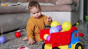 Dark-haired Caucasian child playing with a toy lorry filled with balls. Baby boy pushes the car making sound of a vehicle with his mouth.
