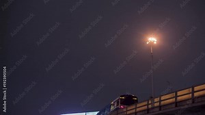 Train moving ahead on overpass bridge against night buildings. Video. Urban landscape on the background, metro train approaching the station on the left. Surface metro station at night.