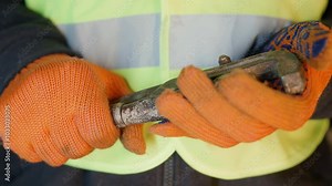 Construction Worker Tapping Pipe Wrench Against Palm: Close-up of Torso in Safety Vest | Impatient Tradesman in Orange Gloves Shows Frustration | Industrial Professional with Tool