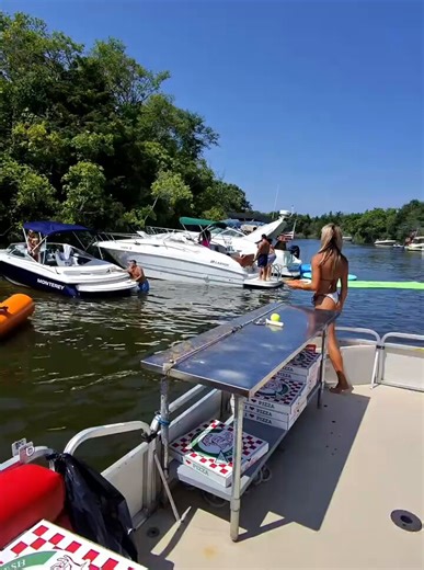 This woman in a white bikini stands on the pizza boat deck surrounded by stacked pizza boxes while a shirtless guy jumps from the boat into the lake.She turns and moves around the stainless steel prep table as other boats and people float nearby under the bright sun with the text Pizza Boat Training in Action on screen.You watch someone run a real floating pizza delivery service on a busy lake day. Do you think it is a brilliant summer hustle that makes the lake more fun or do you say driving a 