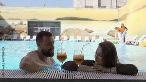 Young muscular man trying to pick up a brunette girl in a swimming pool. Clip. Man flirting with a woman, couple enjoying cocktails.