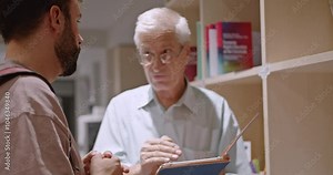 An elderly professor assists a young male student with his schoolwork in the library, using a digital tablet to explain concepts and provide helpful guidance.
