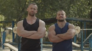 Brutal determined muscular built bodybuilder with well trained bodies standing folded hands at outdoor gym. Confident fit athletic men with arms crossed posing over outdoor sport facilities.