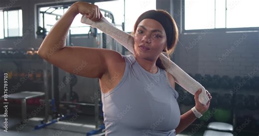 Woman in sleeveless top holding towel behind neck, flexing bicep in gym, checking results on watch