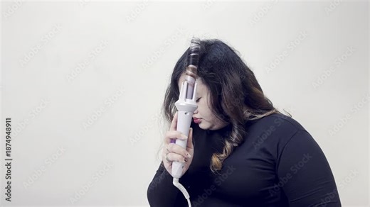 Woman using a curling iron to style her hair with a neutral background, focusing on personal grooming and beauty routines