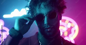 Extravagant man wearing glasses at disco party. Male taking glasses on. Extraordinary man in jacket and silver chain. Glamorous male at neon studio. Man with curly hair at background of LED paintings.