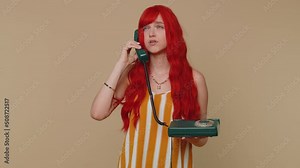 Crazy redhead young woman talking on wired vintage telephone of 80s, fooling, making making playful silly facial faces expressions and grimacing. Red hair ginger girl alone on beige studio background