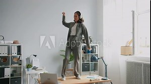 Beautiful office worker in suit is dancing on desk in office wearing headphones enjoying break from work. Crazy people, workplace fun and lifestyle concept.