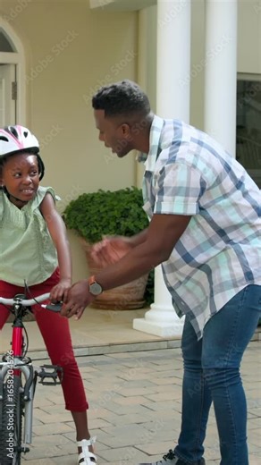 Vertical video: African American dad teaching daughter pedaling and balancing bike in driveway