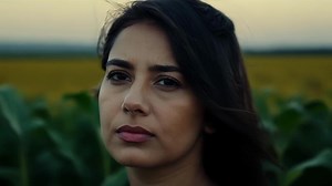 A woman in her thirties her dark hair blowing in the wind as she looks out over the endless rows of corn in the background..