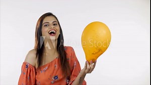 Beautiful Indian girl laughing and bursting a balloon isolated over white background. Young attractive woman popping an orange balloon with a needle and refreshing her childhood memories - leisure ...