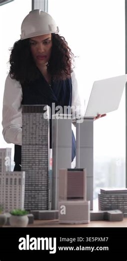 A female architect wearing a hard hat examines building models with a laptop in hand, contemplating urban development Stock Video Footage - Alamy
