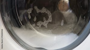 White washing machine washes clothes. Close up footage of spinning drum washing machine. Thick foam, water and bubbles are visible through the glass door of the washing machine. Centrifuge rotation.