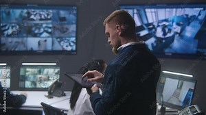Male officer controls security cameras in police surveillance center, uses tablet. PC monitors and multiple big screens on the wall with CCTV cameras footage. Security operators working at background.