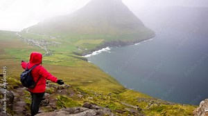 Rear view of person in red rain jacket lifts arms up looking down on Vidareidi village and fjord of Faroe Islands