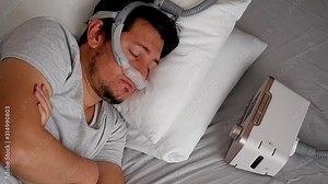 Close-up of healthy young adult man wearing under the nose nasal mask ( CPAP mask ) and using CPAP machine for sleeping smooth without snoring.