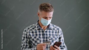 A pleased adult man wearing a protective mask is holding a credit card while using his smartphone standing isolated over a grey background in the studio