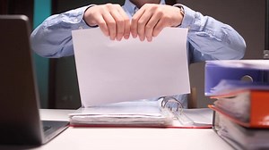 Office worker tearing paper document, contract in half at desk with binders and laptop