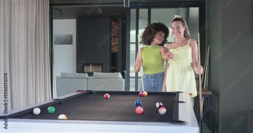 Diverse female friends nudging and embracing at home by black-felt pool table, taking selfies
