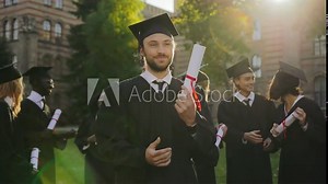 Portrait of the handsome man graduate standing in traditional graduation clothes with diploma in hands and looking to the sky with a hope. Graduates on the background. Outside