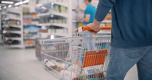 Anonymous Man Pushing a Shopping Cart with Corn Flakes and Other Groceries Through a Modern Supermarket, Navigating Aisles Filled with Various Products and Other Shoppers. Low Angle Follow Footage