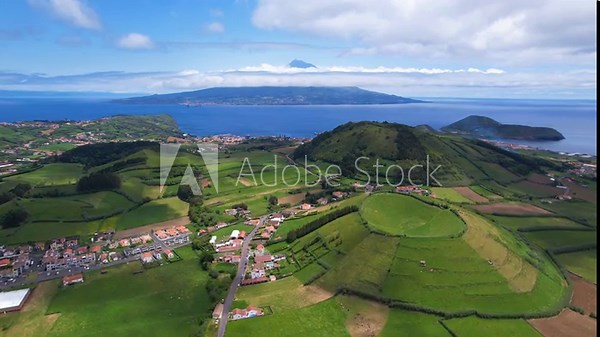 AZORES - 10.27.2025 - Very good aerial footage passing green hills on Faial Island headed towards the water with a view of a Pico Island volcano in the distance.