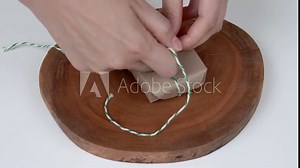 A close-up of a bar of natural soap wrapped in natural brown paper. Woman's hands fold the corners and wrap a string around the parcel, make a bow.
