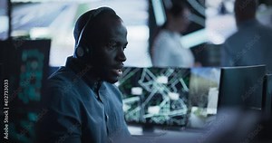 African Specialist in a Dark High-Tech Monitoring Room, Concentrated on a Call and Computer Work. Black Technical Support Manager Using Headphones with Built-In Mic for Communication with Client