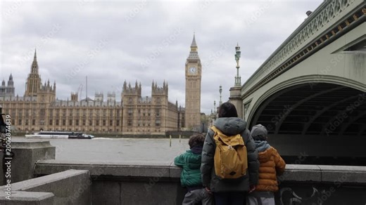 Mother and children watching big ben and the palace of westminster in london from across the river thames on an overcast day with a boat passing under westminster bridge