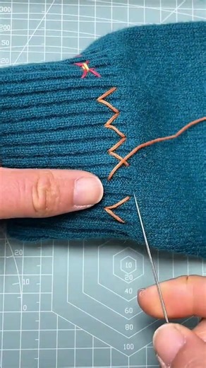 person is hand-stitching a decorative zigzag pattern onto a blue knitted sleeve