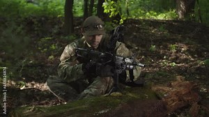 Fully equipped focused military man in camouflage reloading machine gun with ammunition box, cocking weapon shutter and getting ready to shoot while reconnaissance mission in dense forest at daybreak