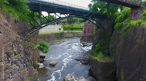 A spectacular 4K drone shot of the historic Paterson Great Falls, located in Paterson, New Jersey. The camera moves forward over the waterfall and under a walking bridge crossing the Passaic River.