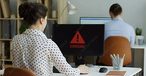 Back view on the angry female office worker coping with a broken computer with an error writing on its screen, shaking it in nerves and male co-worker looking over shoulder. Indoor.