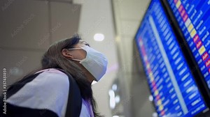 Asian woman looking up at airport schedule time table, wear protective mask, covid-19 pandemic, new normal, flight time schedule, blue led screen, look for check in counter, airport terminal