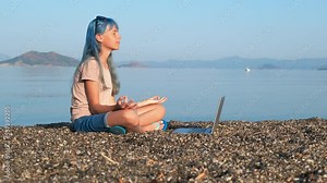 Girl in lotus against sea. A view of pensive girl sits in lotus position by the laptop on the beach against calm blue sea.