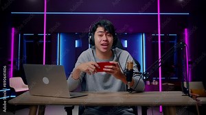 A Smiling Asian Man Gamer With Headphone And Computer Waving Hand And Pointing Index Finger Then Playing Mobile Phone Game In Room Lit By Neon Lights In Retro Stock Video
