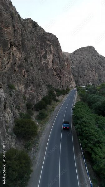 Vertical drone shot along a scenic road between rocky hills and green trees