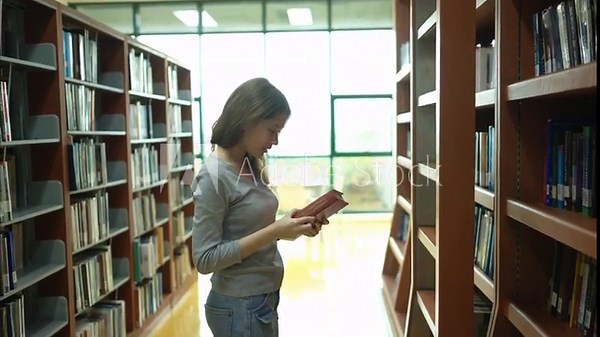 A young woman uses a ladder to pick up a book on the top shelf of a bookcase in a quiet library corridor