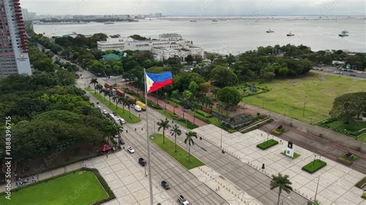 Aerial drone view of iconic Philippine flag flying above Rizal Park's ceremonial plaza with Manila Bay backdrop, colonial buildings, green spaces and vessels anchored in harbor.