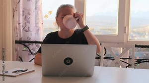 Sleep-deprived student in morning sits at her laptop and drinks coffee, trying to concentrate on writing an abstract article, doing remote work, in home office near window overlooking city.