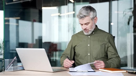 Mature businessman sitting at desk in modern office, turning page on clipboard and signing document, concentrated professional completing paperwork, verifying information and approving business report