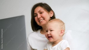 Mom and little daughter are studying at the computer. The laptop is on the table and Mom is teaching her daughter how to use the computer. People in white shirts against the background of white walls