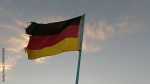 Germany flag on flagpole flutters in the wind at sunset, slow motion, A dirty and tattered flag of the Federal Republic of Germany on blue sky with clouds background