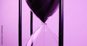 Closeup of sand timer clock with sand flowing from through hourglass on purple tone.